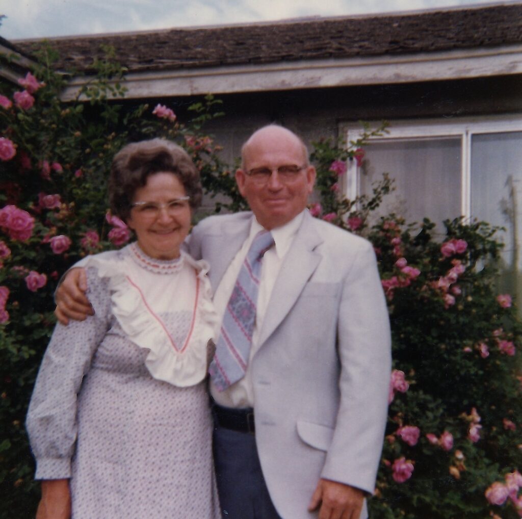 Grandparents standing in front of a blooming rosebush. 
